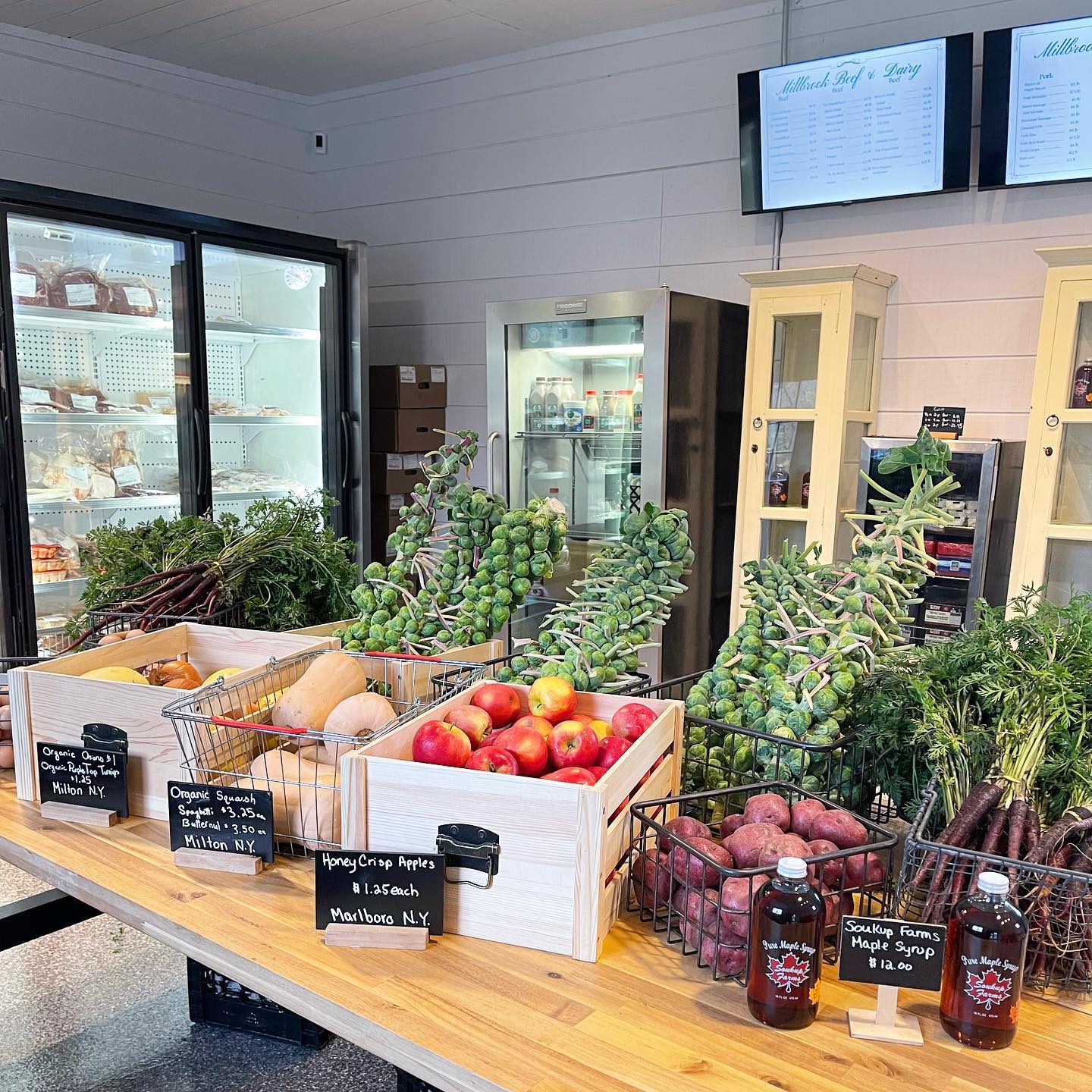 Fresh produce display featuring Honeycrisp apples, organic squash, and seasonal vegetables in a local market. Baskets filled with colorful carrots, Brussels sprouts, and potatoes showcase farm-to-table offerings. Refrigerated section visible in the background with dairy products.