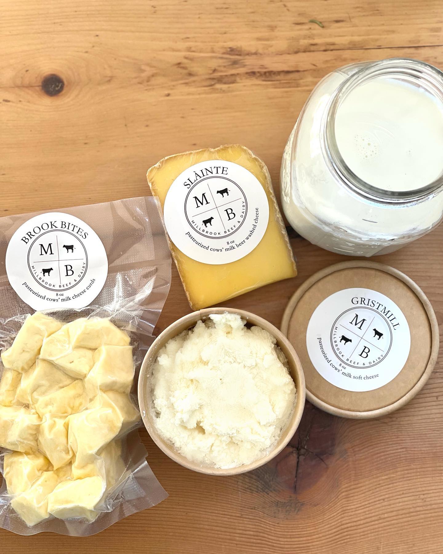 Assorted dairy products from Millbrook Beef & Dairy, including Brook Bites cheese curds, Slainte aged cheese, and Gristmill soft cheese, alongside a jar of fresh milk, arranged on a wooden surface.