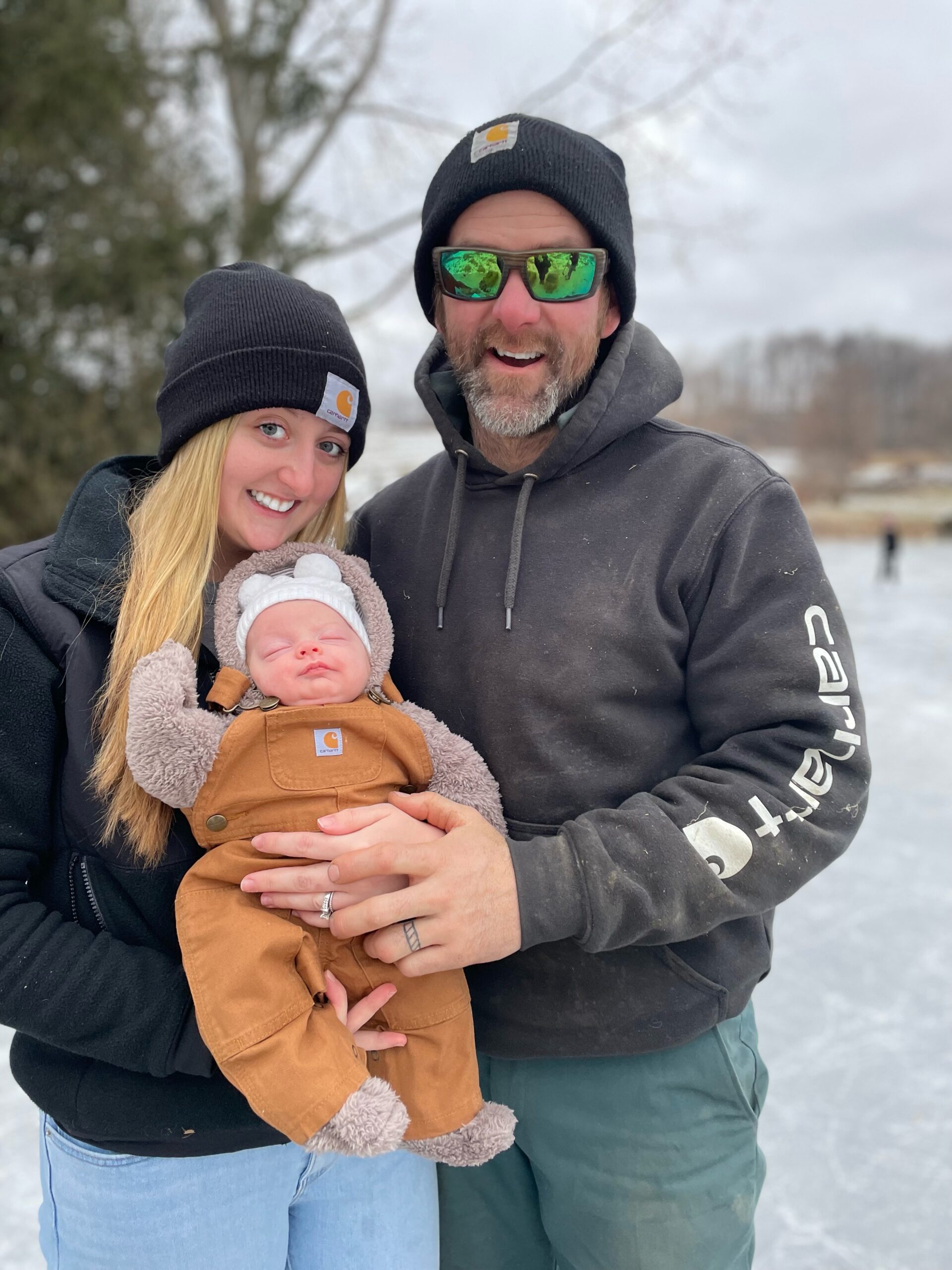 Family portrait on an ice rink featuring a couple wearing matching black beanies and a baby in a cozy brown snowsuit, showcasing winter fun and family bonding in a snowy outdoor setting.