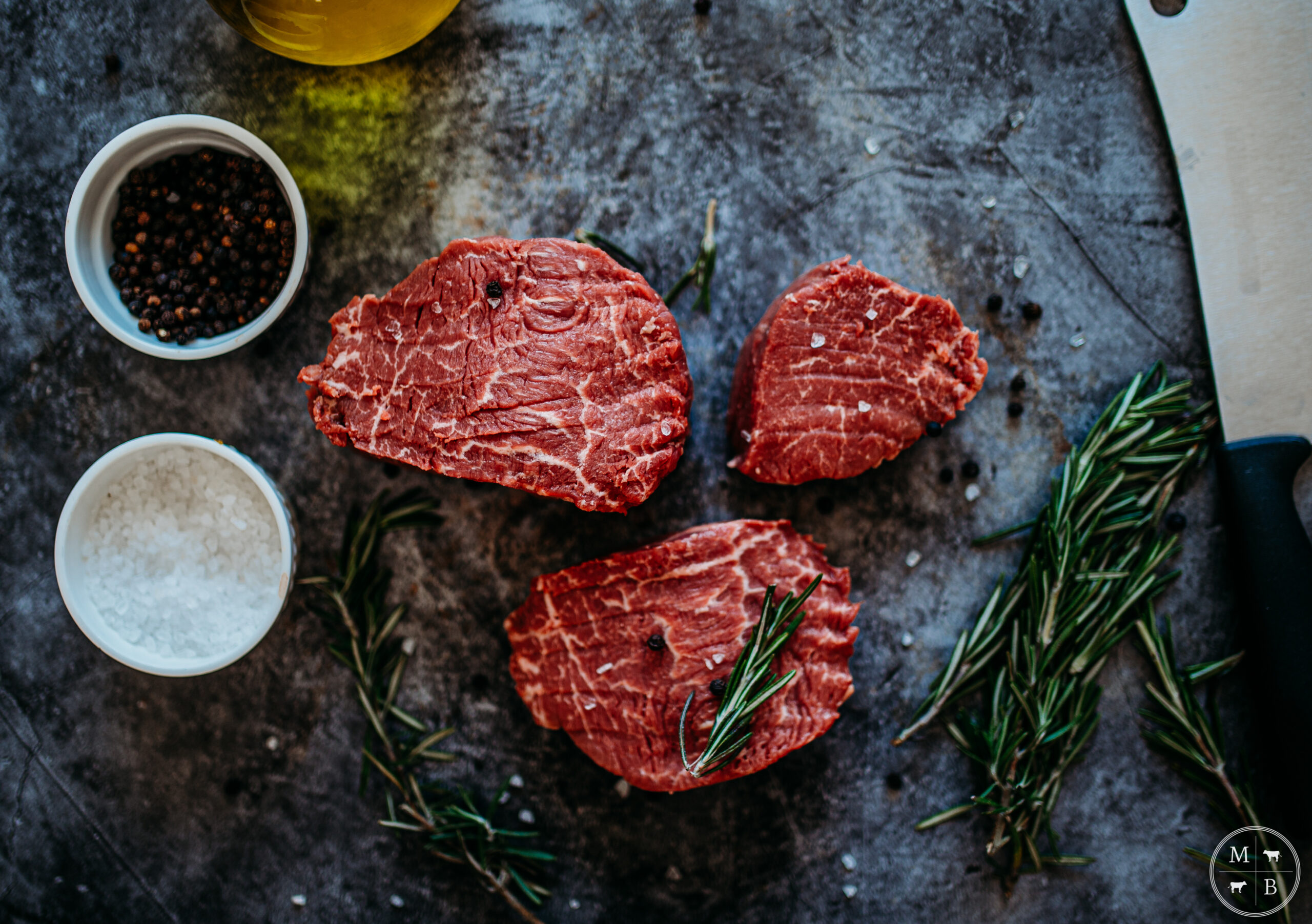 Raw beef cuts arranged on a rustic surface with rosemary sprigs, black peppercorns, and sea salt in small bowls, highlighting fresh ingredients for cooking.