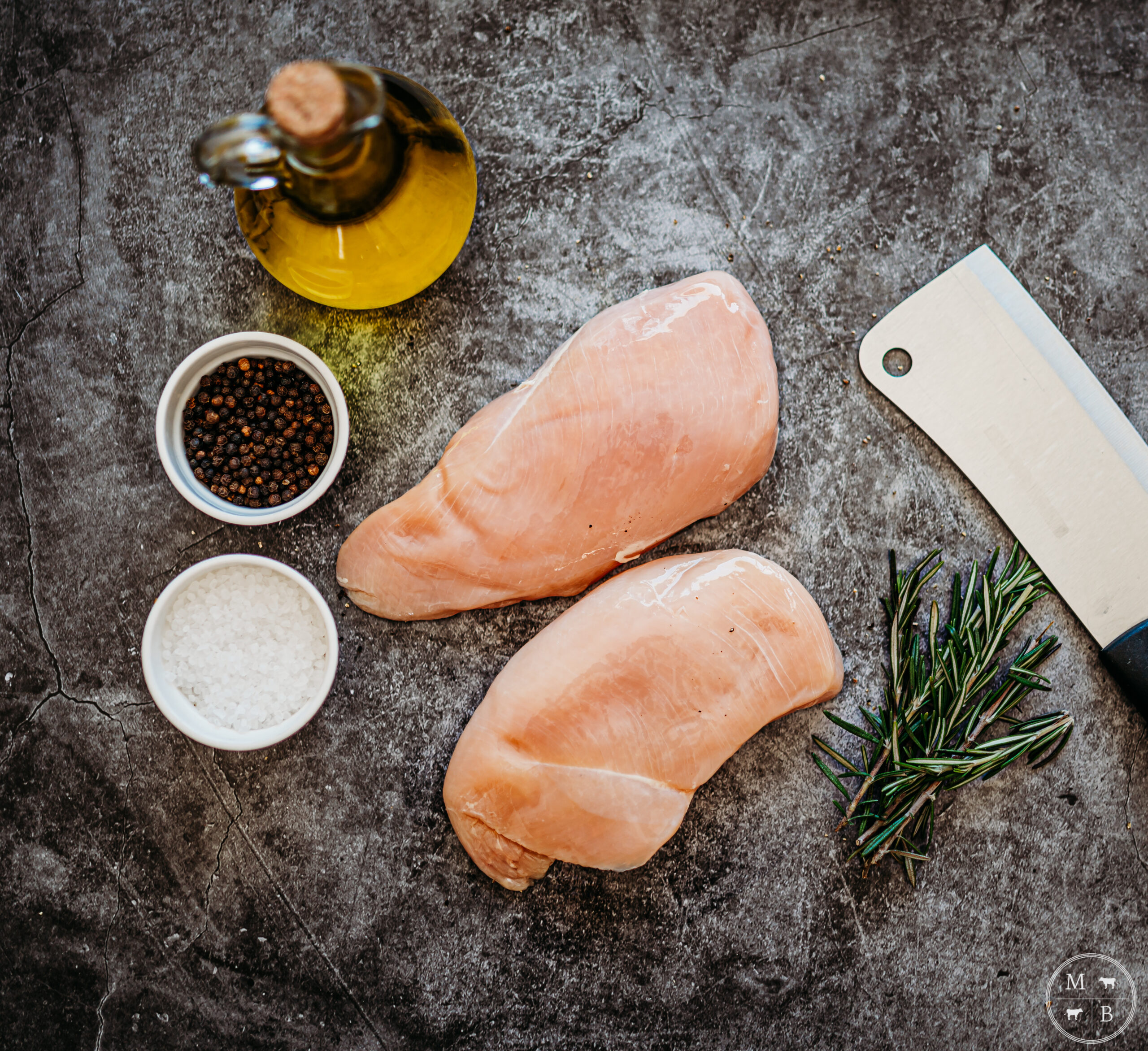 Raw chicken breasts, olive oil, black peppercorns, sea salt, fresh rosemary, and a kitchen knife arranged on a textured surface, ready for cooking.