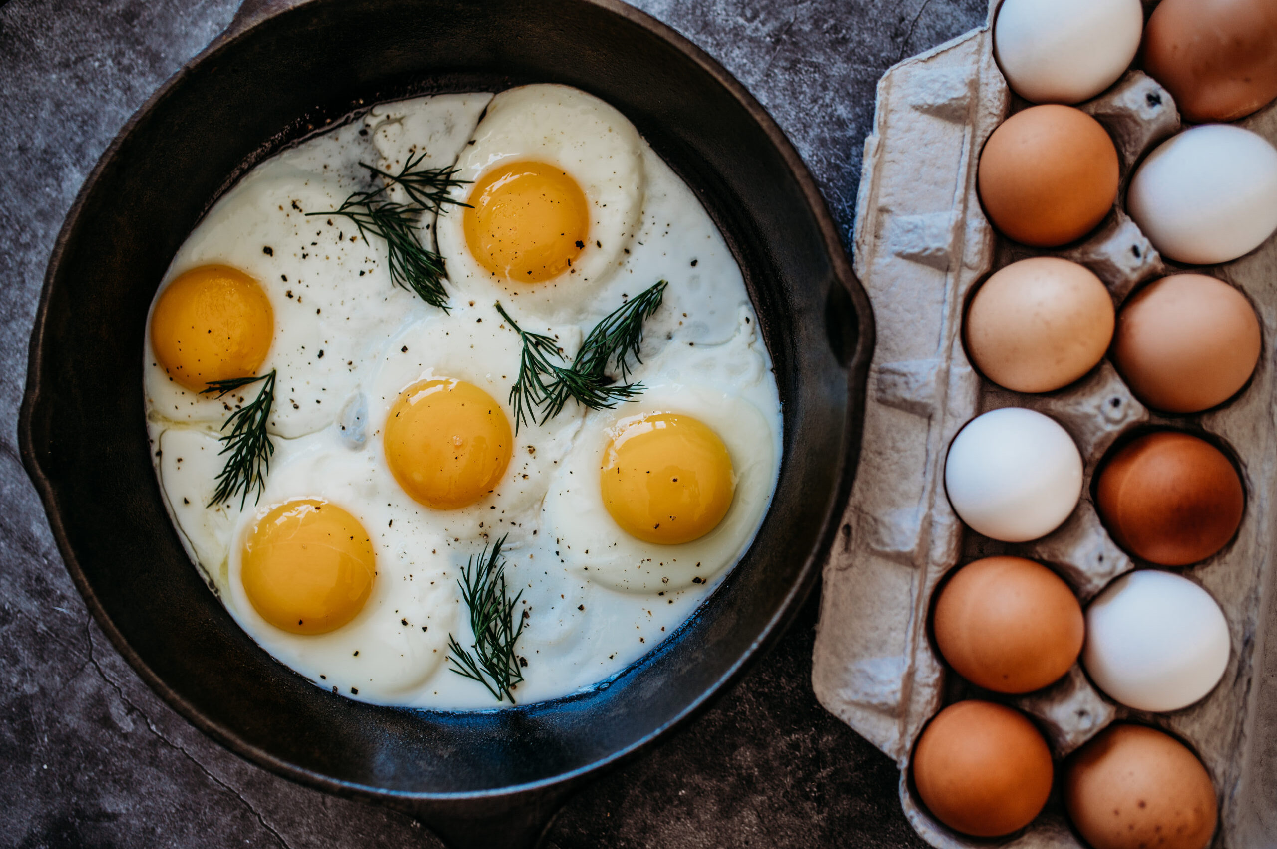 Fried eggs with bright yellow yolks cooked in a cast iron skillet, garnished with fresh dill and black pepper, alongside a carton of assorted brown and white eggs.