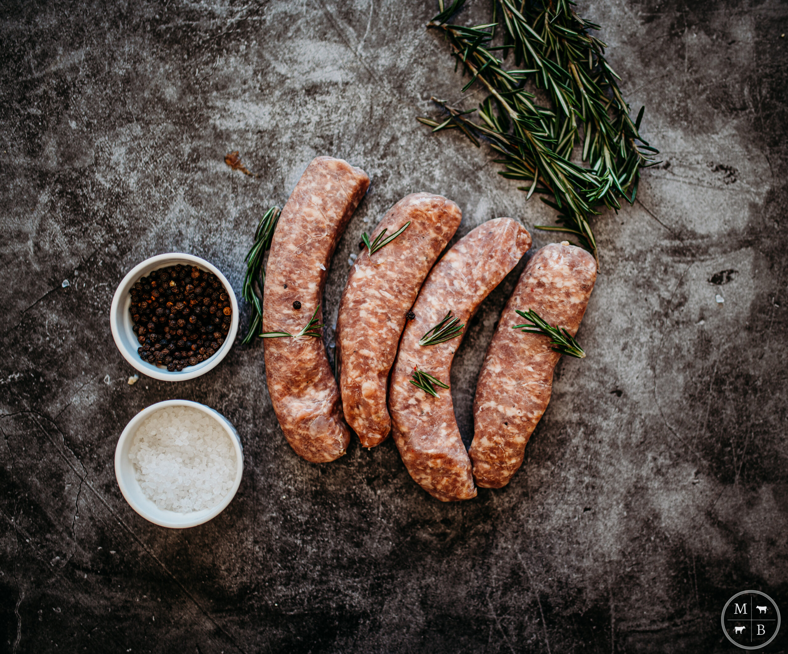 Raw sausages arranged on a textured surface, accompanied by bowls of black peppercorns and coarse sea salt, garnished with fresh rosemary sprigs for flavor enhancement.