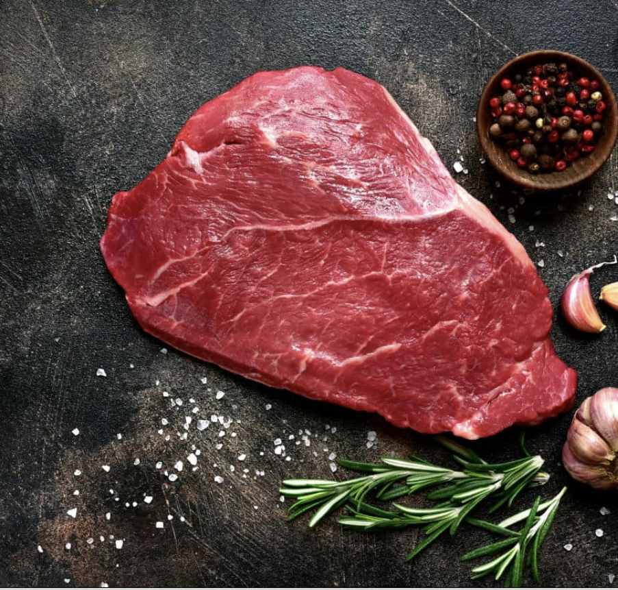 Fresh, raw beef steak on a dark slate surface, accompanied by garlic cloves, rosemary sprigs, and a bowl of mixed peppercorns, showcasing ingredients ideal for flavorful cooking.